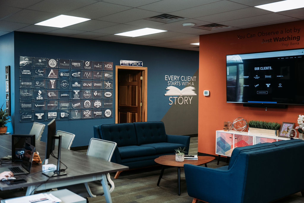 View of the 898 office featuring client logos, desks, laptops, televisions, couches, and a quote that says "Every Client Starts with a Story" and an open book in the center of the quote.