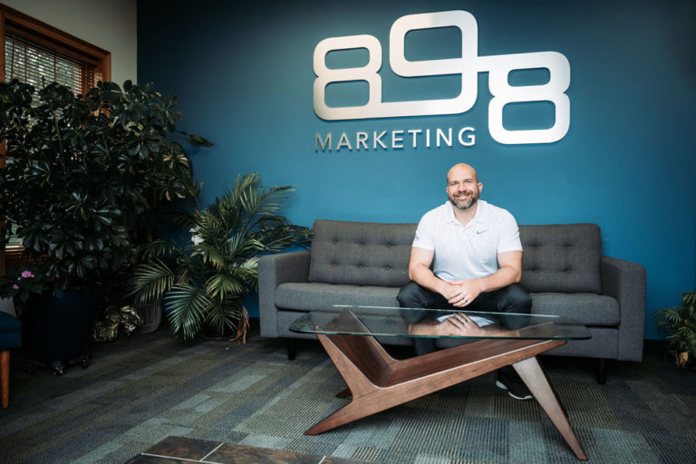 Jeff Ryznar sitting on a couch in front of the silver 898 logo on a blue wall.